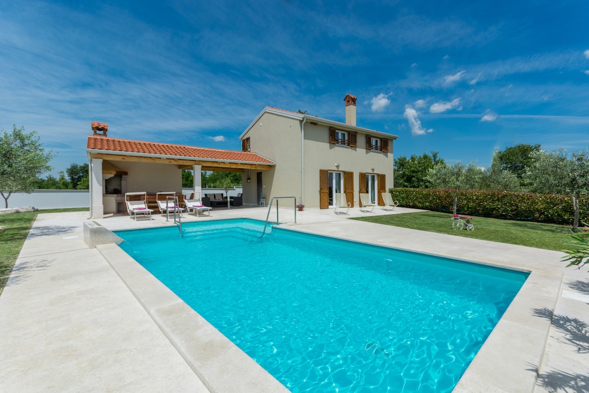 The image features a modern detached house with a red-tiled roof, framed by a spacious outdoor area. A clear swimming pool is visible, complemented by a comfortable terrace housing a covered outdoor kitchen and lounge chairs. Lush greenery surrounds the property, creating a tranquil environment.