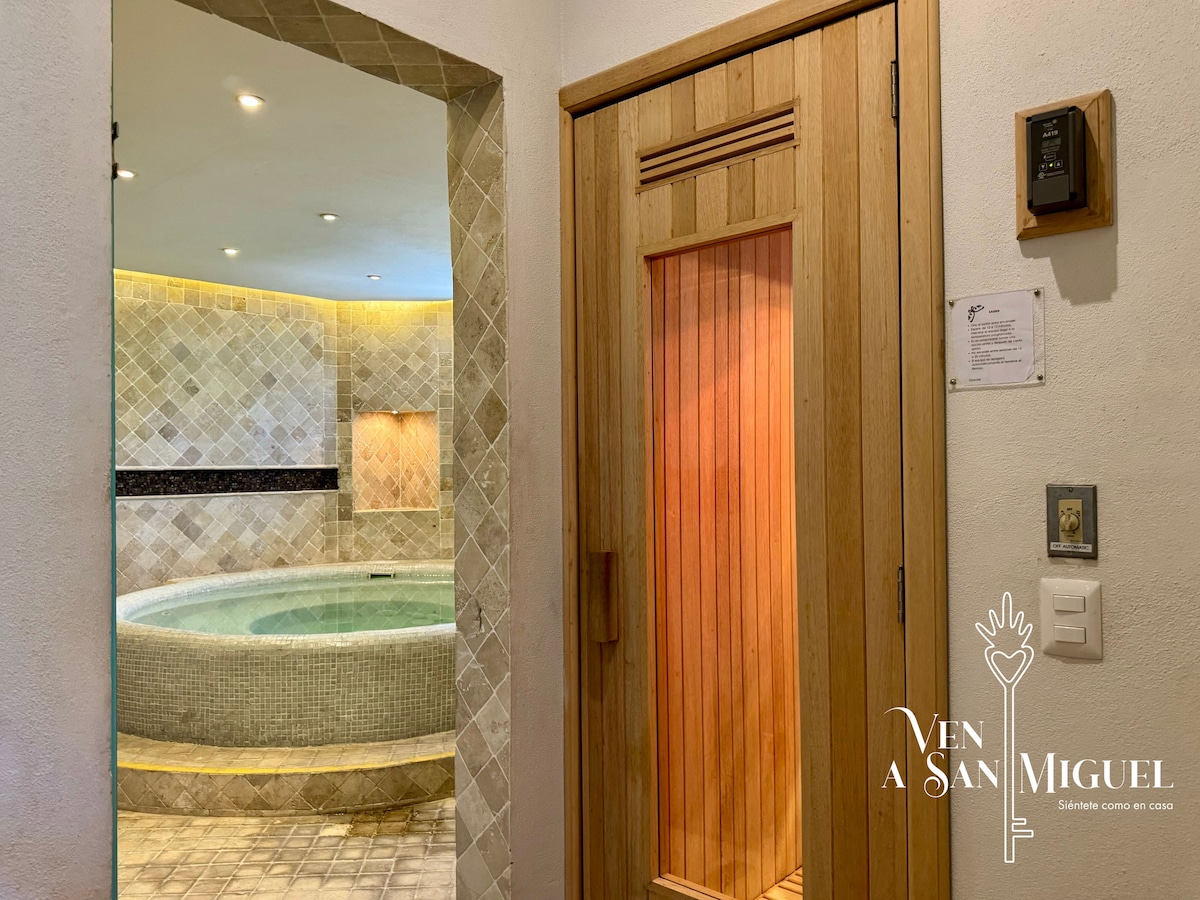 An inviting wellness area features a wooden door leading to a sauna, adjacent to a circular jacuzzi. Textured stone tile surrounds the jacuzzi, providing a calming environment. Soft lighting enhances the serene atmosphere for relaxation and rejuvenation.