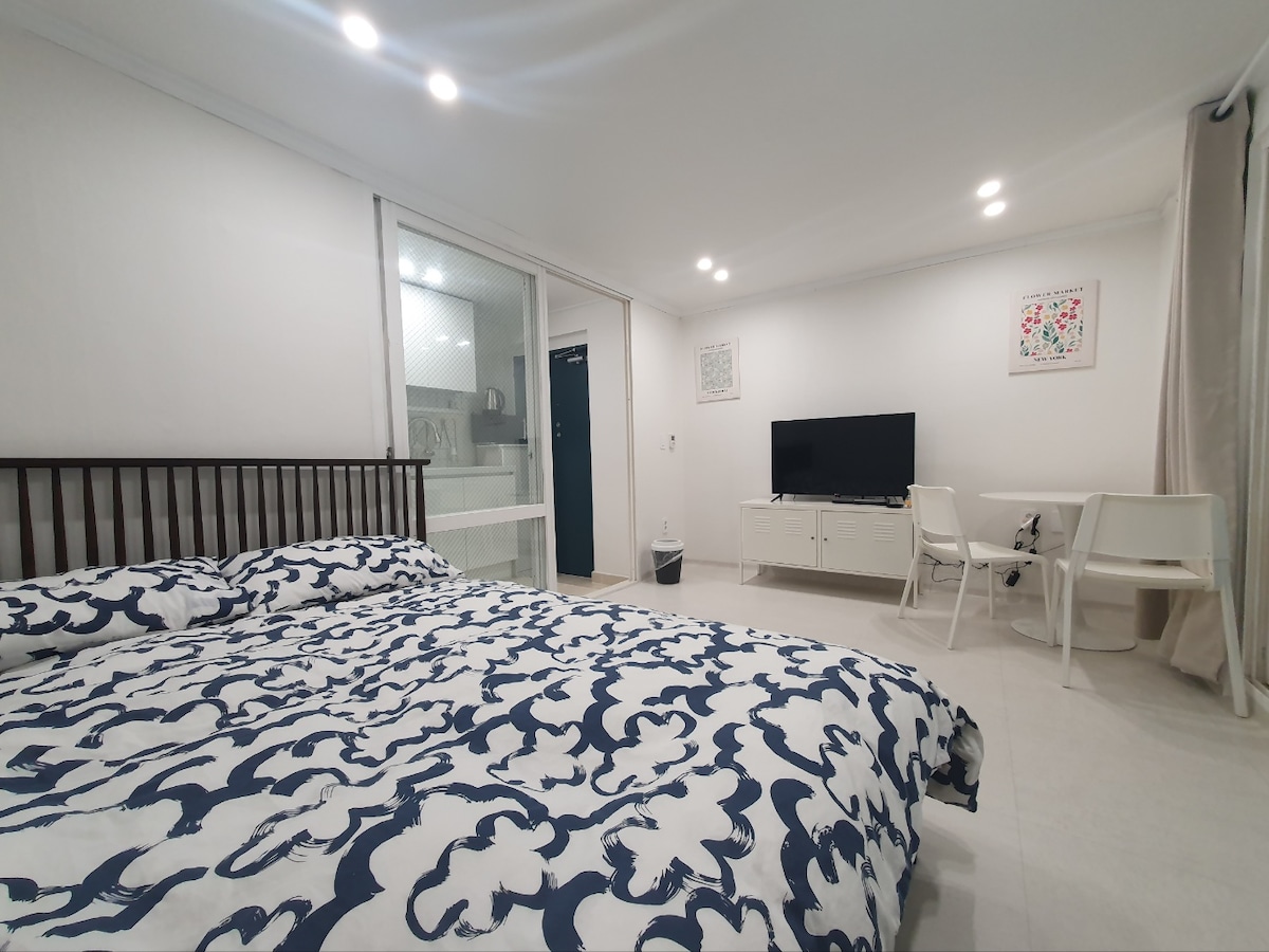 A spacious sleeping area features a large bed covered with a patterned comforter. A television is mounted on the wall opposite the bed, accompanied by a white media console. Nearby, two chairs and a small table are positioned, creating a cozy space for dining or relaxation.