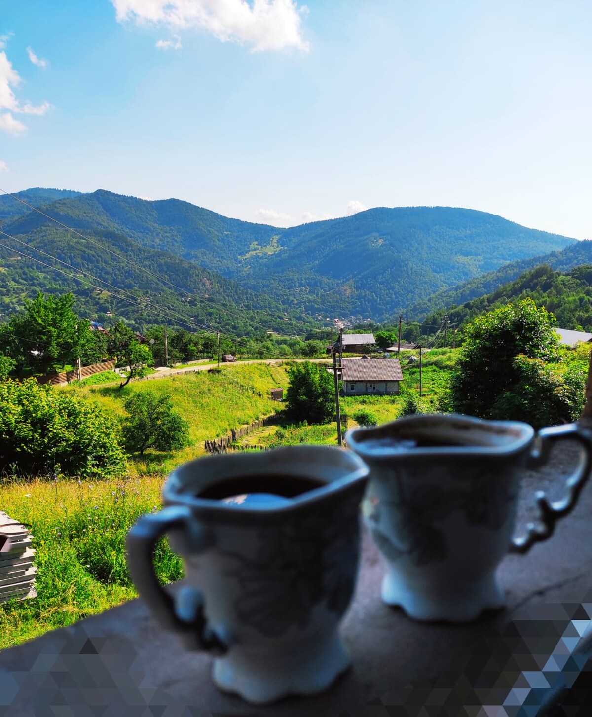 A scenic mountain view is presented through a window, with rolling hills and a winding road visible. Two delicate cups of coffee are placed in the foreground, drawing attention to the lush greenery and distant houses nestled among the hills.