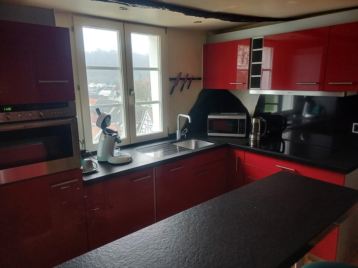 A modern kitchen showcases sleek red cabinets and a dark countertop. Appliances include an oven, microwave, and coffee maker. Natural light filters through a window, illuminating the space. A bar countertop for two complements the kitchen layout, providing a functional and stylish environment.