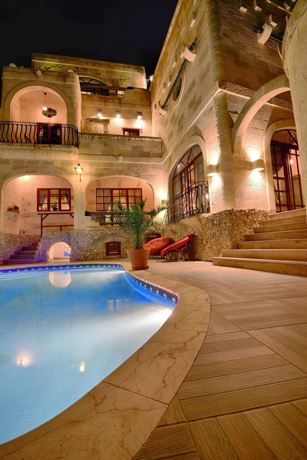 A well-lit courtyard features a serene pool, reflecting soft illumination from surrounding architecture. Steps lead up to areas adorned with warm stone walls and multiple arched windows. Comfortable lounge chairs are positioned at the pool's edge, inviting relaxation.