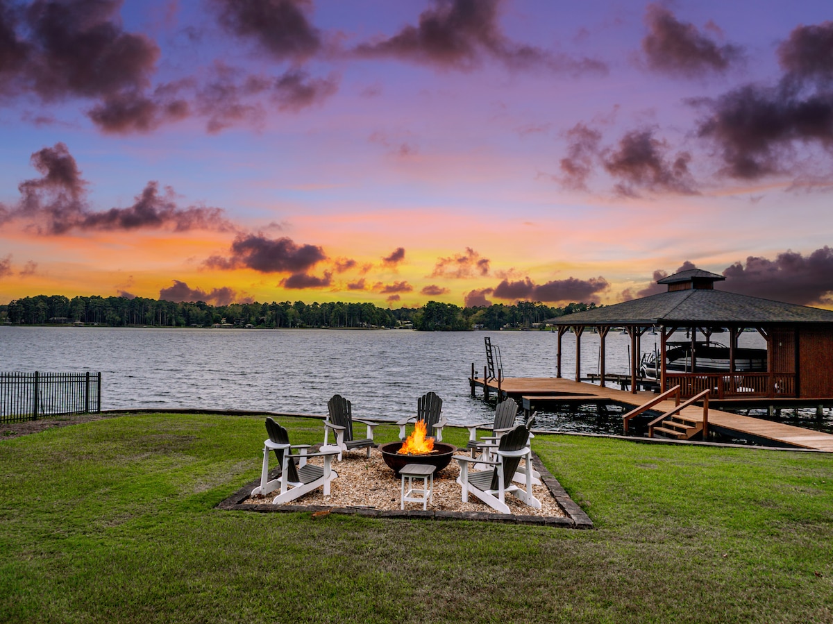 A fire pit surrounded by six white Adirondack chairs is positioned in a manicured lawn, overlooking a tranquil lake. A gazebo is visible on the right, with vibrant sunset colors reflected in the sky, enhancing the serene lakeside setting.