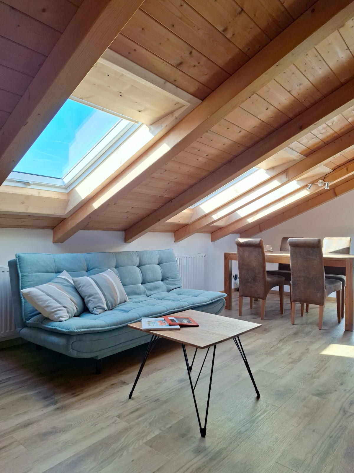 A light-filled living area features a comfortable blue sofa and a small coffee table, complemented by two large skylights and wooden beams overhead. A dining table with four chairs is situated nearby, highlighting the open and airy feel of the space.