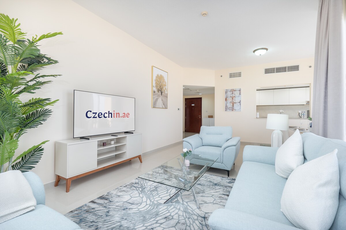 A light-filled living area features modern furnishings in soft blue tones. A sleek television stands on a white console, complemented by a marble-patterned area rug. Two comfortable armchairs are positioned around a small table. The open kitchen is visible in the background, enhancing the spaciousness.