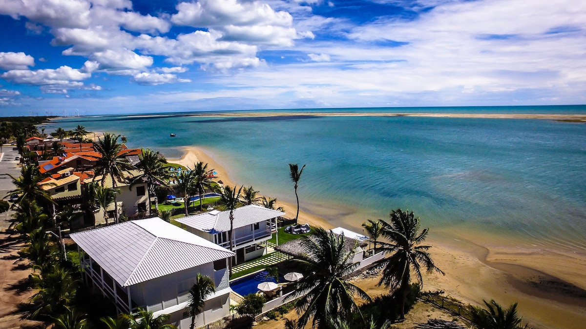 An aerial view captures the beachfront property adjacent to a serene lagoon. Lush palms frame the scene, with clear turquoise waters extending toward the horizon. The white house features a spacious patio and swimming pool, all set against a backdrop of sandy coastline and distant landforms.