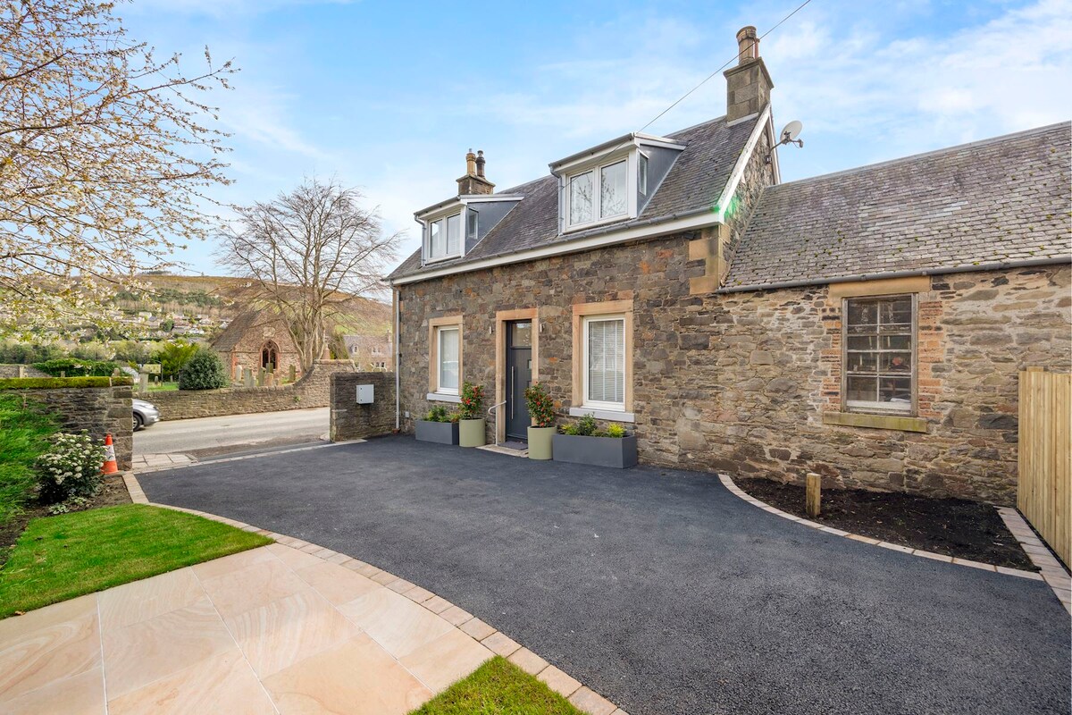 A newly renovated cottage exterior is displayed, featuring a stone facade with large windows and a well-maintained garden. A private off-street parking area is evident, alongside planters with greenery flanking the entrance. The surrounding landscape includes rolling hills under a bright sky.