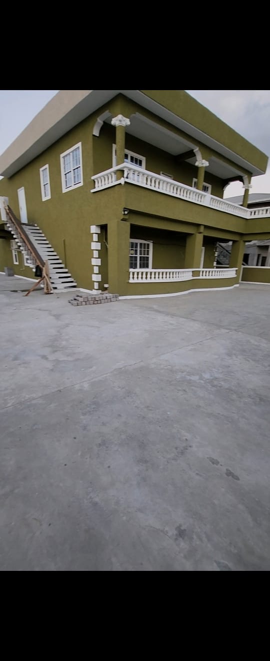 A two-story structure is presented with a distinctive green exterior and white railings. A staircase leads to the upper level, while the spacious concrete area in front offers ample space for family activities. Windows allow natural light to brighten the property.