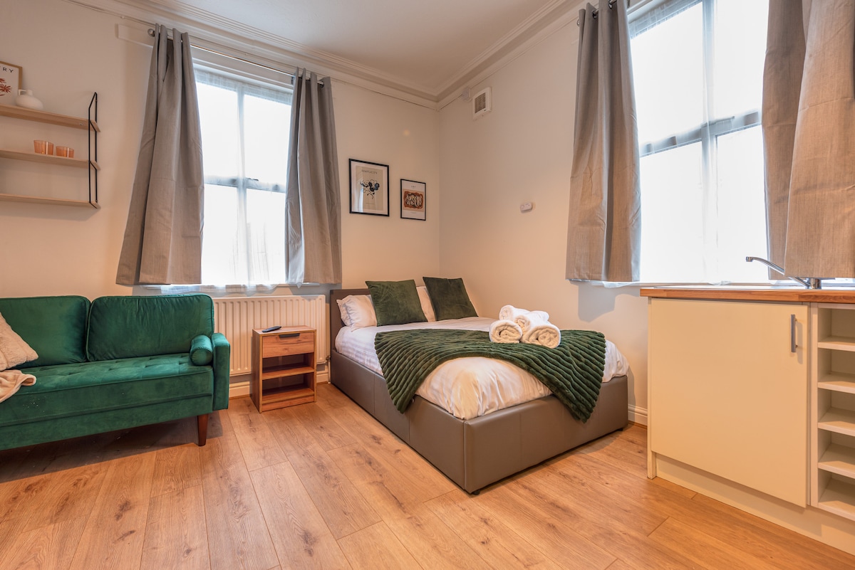 A modern studio flat features a spacious bed with a dark green throw. Natural light enters through two large windows adorned with light curtains. A comfortable green sofa is positioned nearby, alongside a small wooden nightstand and a kitchen area with essential appliances.