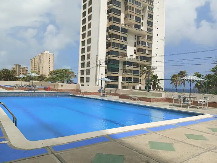 A large swimming pool is shown surrounded by a spacious deck. Sun loungers and umbrellas are positioned around the pool area. In the background, a tall residential building stands alongside palm trees, with the ocean visible in the distance under a partly cloudy sky.