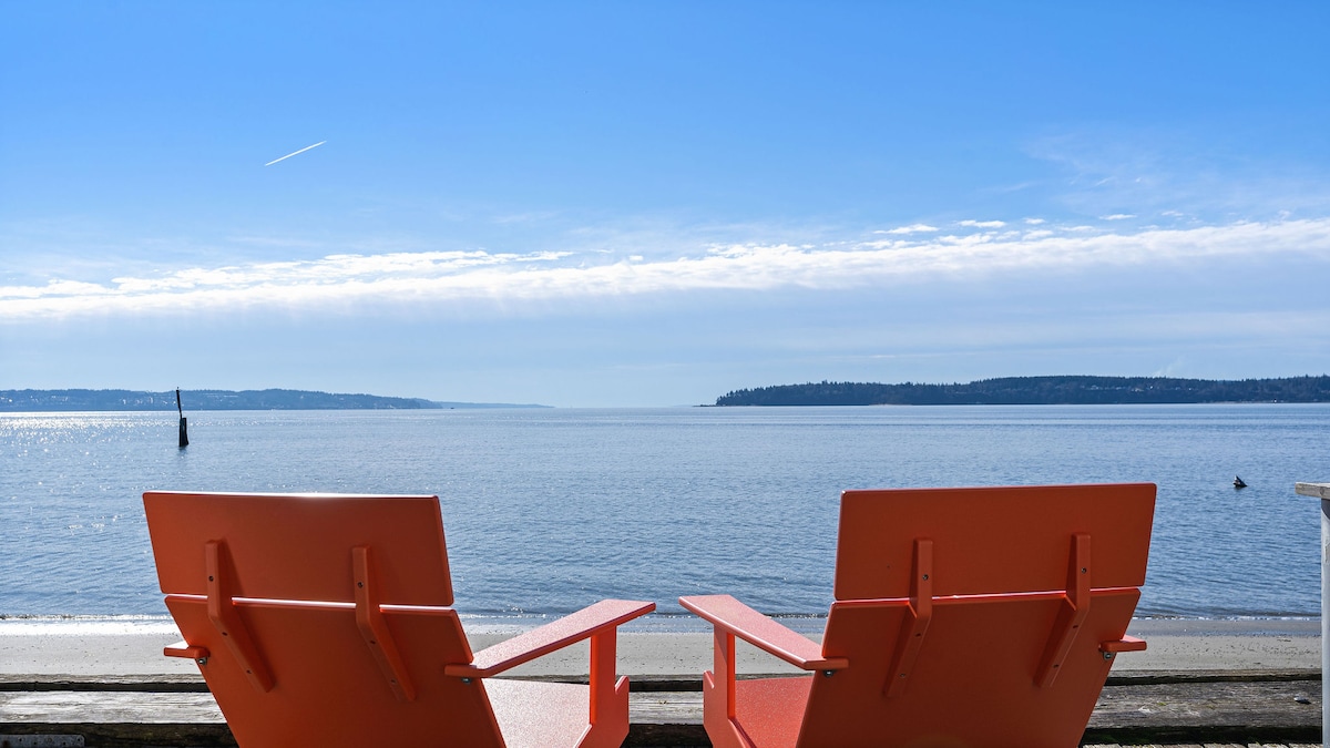 Two bright orange chairs are positioned on the sandy beach, facing the calm water. The view stretches across the sound, with gentle waves rippling under a clear blue sky, creating a serene and inviting coastal scene.
