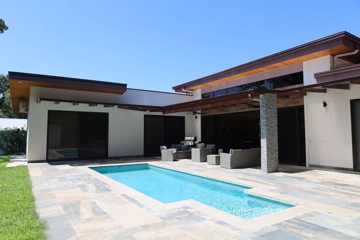 The outdoor area features a secluded private pool surrounded by stone tiles, with comfortable seating arrangements positioned nearby. Large glass doors connect the interior with the exterior, allowing natural light to fill the space. Lush green grass surrounds the pool, enhancing the tranquil setting.