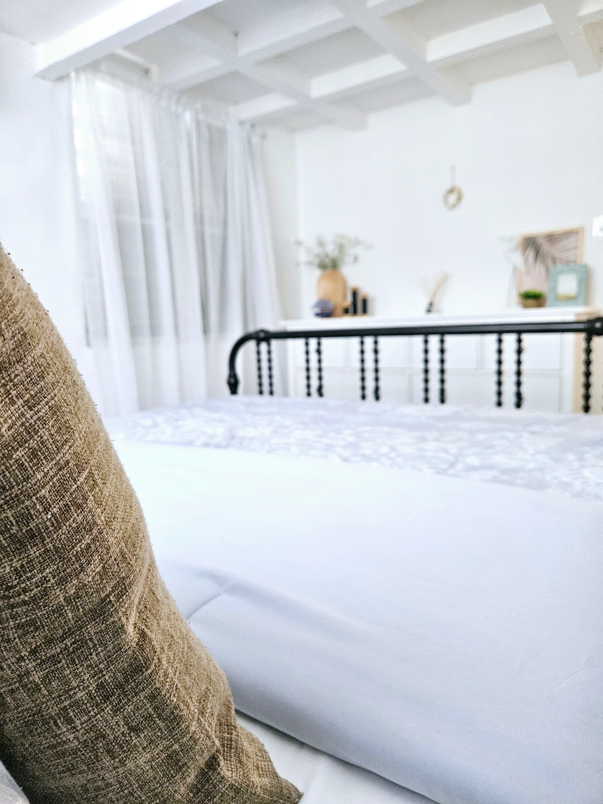 A cozy sleeping area is featured, showcasing a spacious bed adorned with soft linens. Natural light filters through sheer curtains, illuminating a decorated nightstand in the background. The room's white walls and wooden beam ceiling contribute to a serene atmosphere.
