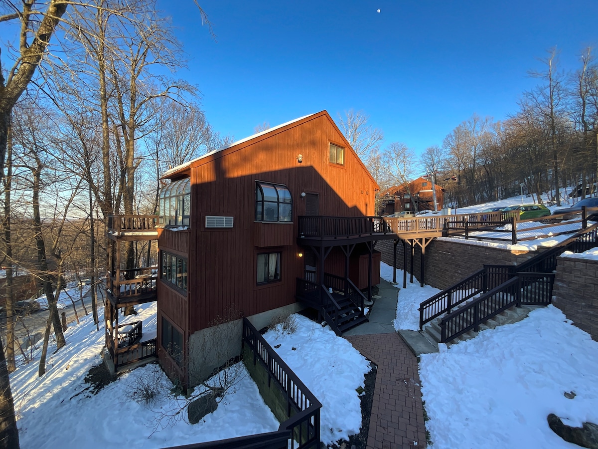 The exterior of a wooden cabin is shown, featuring multiple balconies with wooden railings. Snow covers the ground and pathways, while tree branches are bare. A clear blue sky and a moon can be seen above, highlighting the peaceful surroundings.