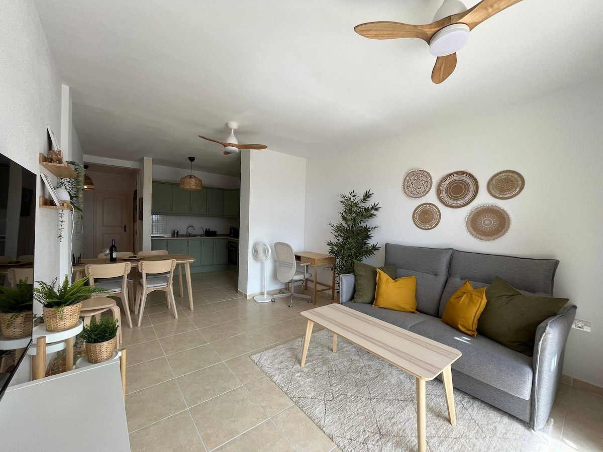 A spacious living area features a comfortable gray couch adorned with pillows in shades of yellow and green. A light wooden coffee table is centered on a neutral area rug, with a fan overhead. Nearby, a dining table and small workspace are visible, enhancing the inviting atmosphere.