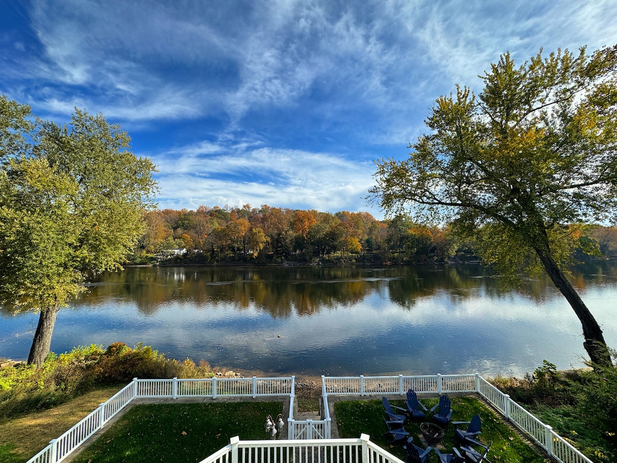 A serene river view is presented, showcasing calm waters reflecting a blue sky with fluffy clouds. Colorful trees line the opposite bank, hinting at autumn hues. In the foreground, a white fence encloses a grassy area, with a seating arrangement visible beside the water.