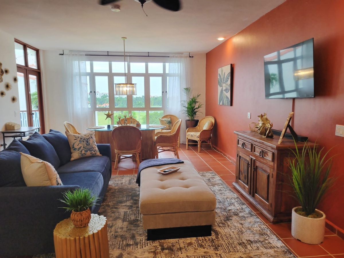A spacious living area features a comfortable dark sofa and a stylish tufted ottoman. Light floods in from large windows, illuminating the neutral-toned decor and highlighting a wooden sideboard. A dining table with rattan chairs completes the inviting arrangement, all set against a warm accent wall.