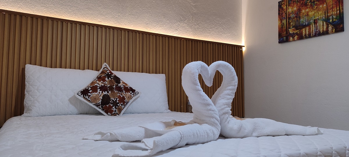 A cozy sleeping area is described, featuring a spacious bed adorned with white bedding. Two towel swans arranged in a heart shape are displayed at the foot of the bed. A decorative pillow adds color against the wooden accent wall, with soft lighting enhancing the room's ambiance.