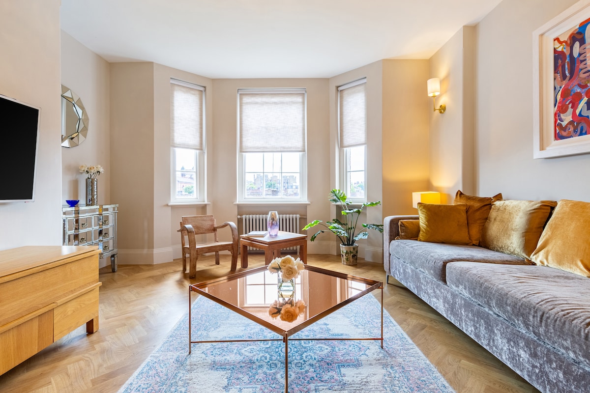 A spacious living area is furnished with a plush sofa, wooden coffee table, and accent chairs. Natural light fills the room through large windows, highlighting the warm tones of the decor and the intricate pattern of the area rug.