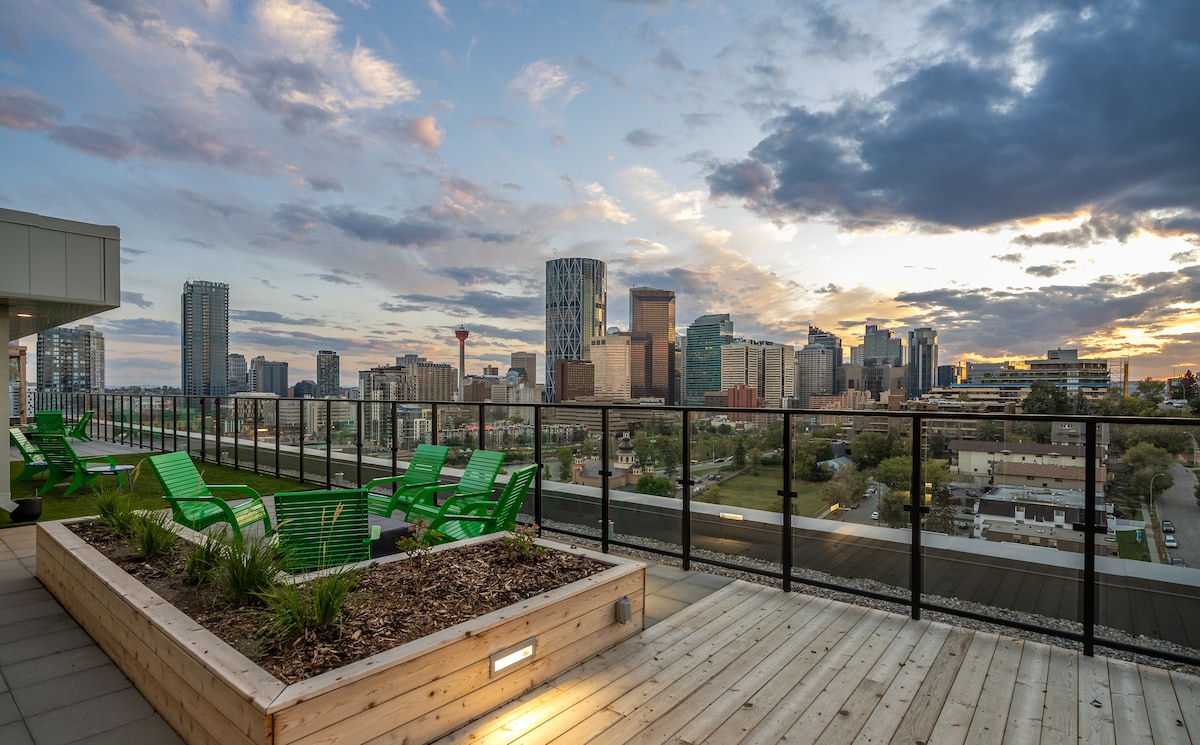 A spacious rooftop terrace offers sweeping views of Downtown Calgary, showcasing a blend of modern skyscrapers and natural landscapes. Green lounge chairs are arranged thoughtfully around landscaped planters, providing a comfortable setting to enjoy the sunset and city skyline.