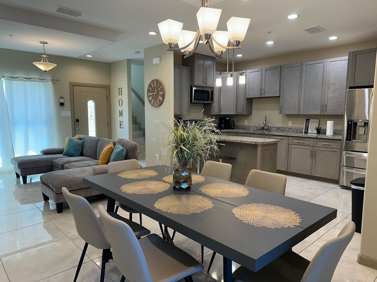 A spacious dining area features a large black table surrounded by light-colored chairs. In the background, a modern kitchen with stainless steel appliances and granite countertops is visible, complemented by a cozy gray sofa and natural light from nearby windows.