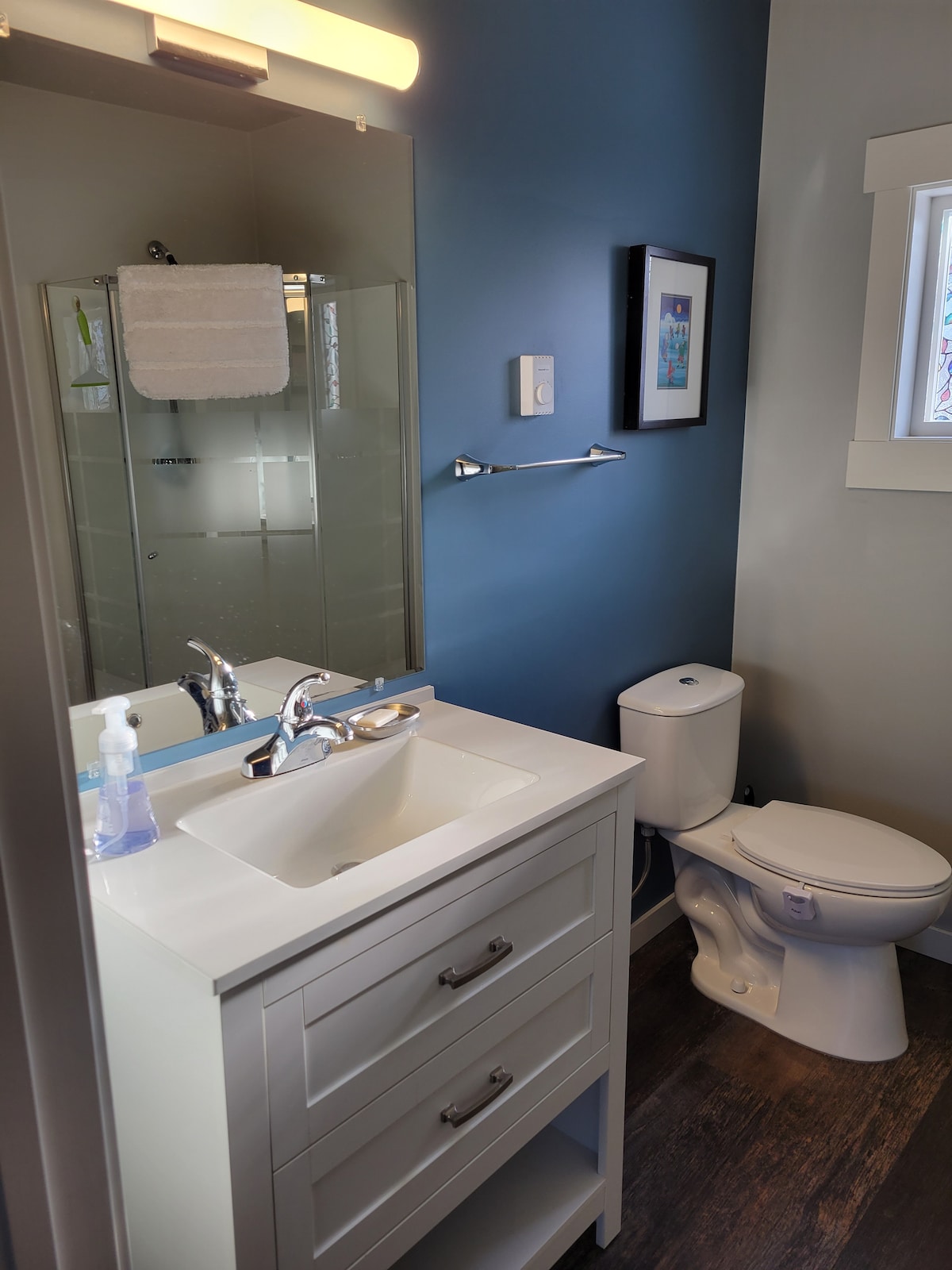 A modern bathroom is presented with light grey cabinetry and a sleek sink. A glass shower is visible in the corner, while a white towel hangs neatly on a rack. The walls are painted in a calming blue, complemented by a small framed artwork.