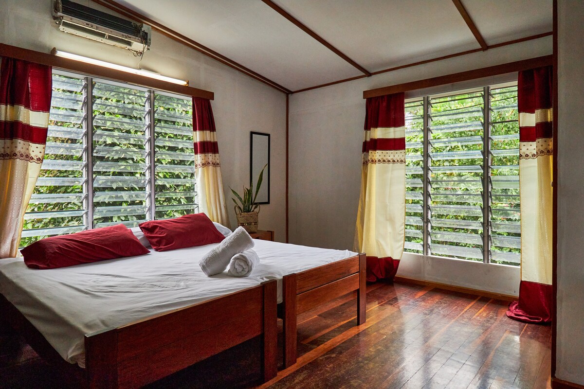 A spacious bedroom features large windows adorned with light-colored curtains, allowing natural light to stream in. A wooden bed with red accent pillows is centrally positioned. The room’s wooden flooring adds warmth, complemented by the presence of a green plant in the corner.
