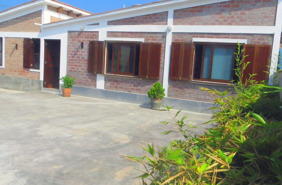 The exterior of the home features a modern design with brick walls and wooden shutters. A spacious patio area is visible, along with potted plants adding greenery to the setting. The entrance door is framed by a welcoming pathway.