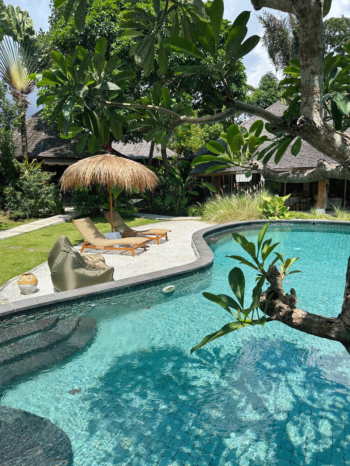 A tranquil pool area is framed by lush greenery and features a thatched umbrella shading a lounge chair. The clear water reflects sunlight, while the stone pathway curves around the pool, leading to a nearby garden. Tropical plants enhance the serene atmosphere.