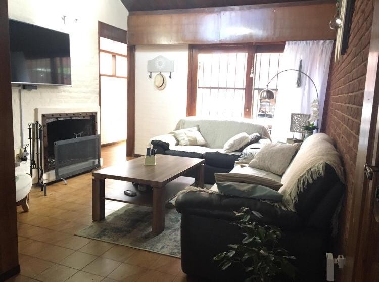 A cozy living room features a large coffee table at its center, surrounded by a mix of sofas and armchairs. A wall-mounted television is installed beside a fireplace. Natural light enters through several windows, highlighting the textured wall and warm color palette.