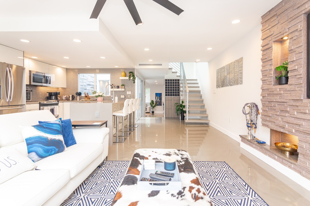 An open-concept living area features a modern white sofa adjacent to a stylish coffee table placed on a geometric rug. The space is illuminated by recessed lighting, with a view of the kitchen and dining area adorned with sleek finishes. A staircase and decorative wall accents enhance the contemporary design.