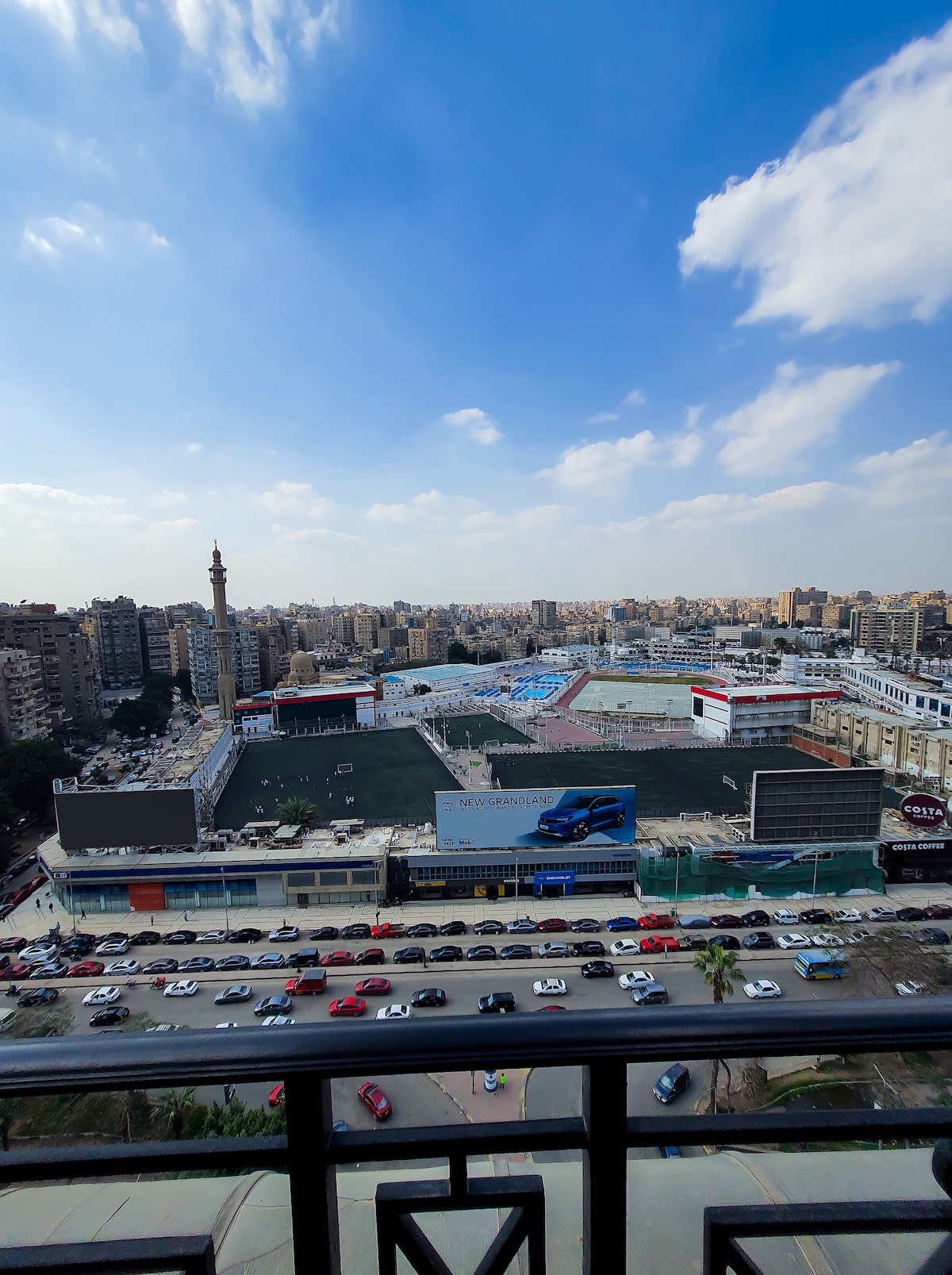 A panoramic view from the balcony showcases the bustling scene of University Street. High-rise buildings and busy streets are visible, alongside a large sports complex. The blue sky with scattered clouds adds to the vibrant city atmosphere.