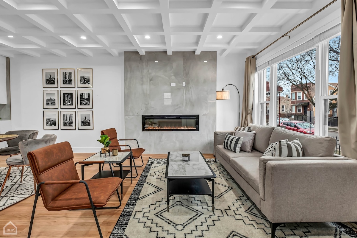 A modern living area features a contemporary design with a stylish gray sofa and brown leather chairs. A sleek, geometric area rug sits beneath a coffee table, while framed black-and-white photographs line the walls. Large windows allow natural light to illuminate the space.