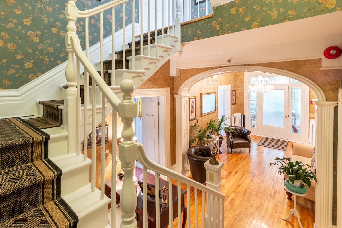 An inviting staircase with a decorative handrail leads to a second level. The entrance area features hardwood floors and a floral wallpaper backdrop. Comfortable seating arrangements are present in the lobby, complemented by plants and a chandelier, enhancing the open space.