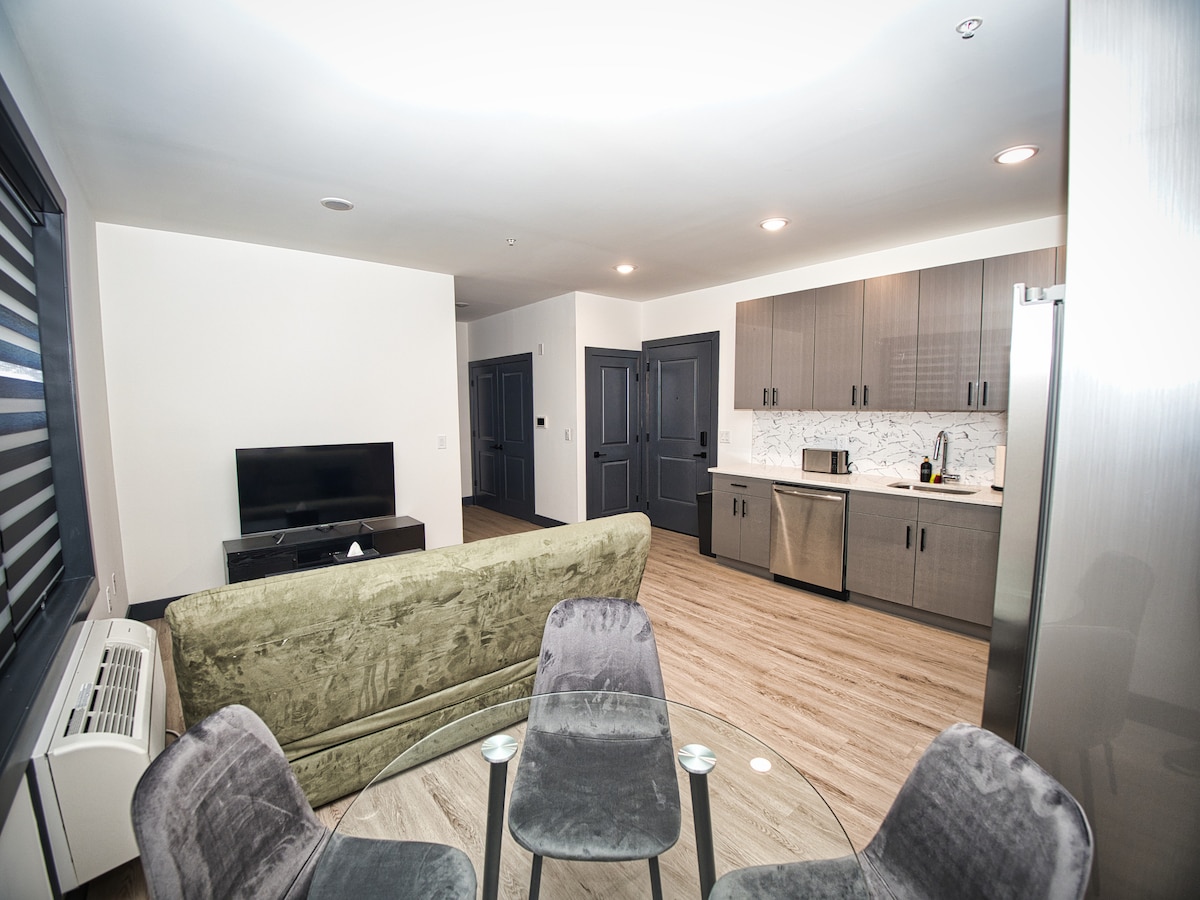 An open living space features a modern kitchen with gray cabinetry and marble countertops, alongside a dining table with four chairs. A green couch is positioned in the foreground, with a television visible against the wall. Warm overhead lights enhance the inviting ambiance.
