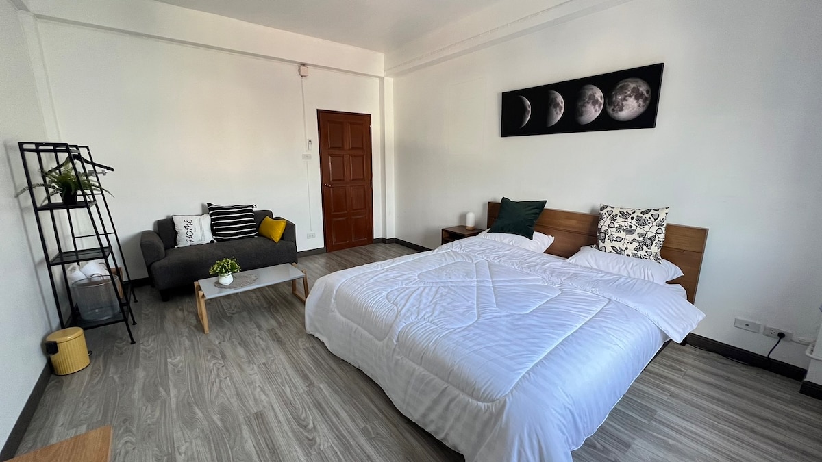 A spacious sleeping area features two beds with crisp white linens. A gray couch with decorative cushions is positioned against the wall. A small table and a plant stand add a touch of greenery, while a moon-themed artwork decorates the wall above.