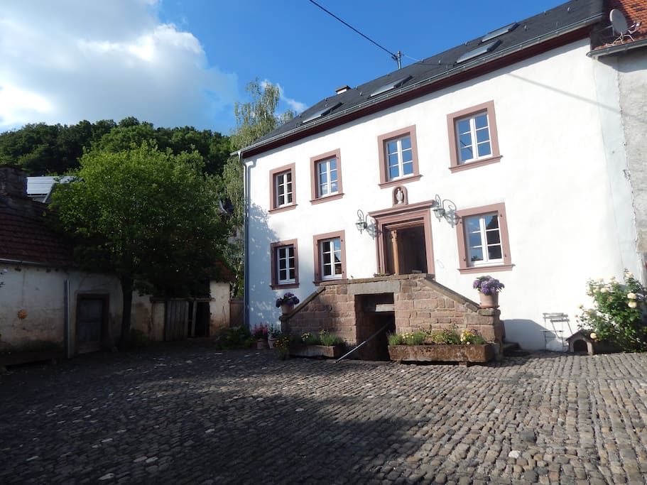 The exterior of a historic building is shown, featuring a symmetrical façade with large windows and a central entrance accentuated by steps. Floral arrangements adorn the entryway, while cobblestone ground and greenery surround the structure, creating a charming courtyard atmosphere.