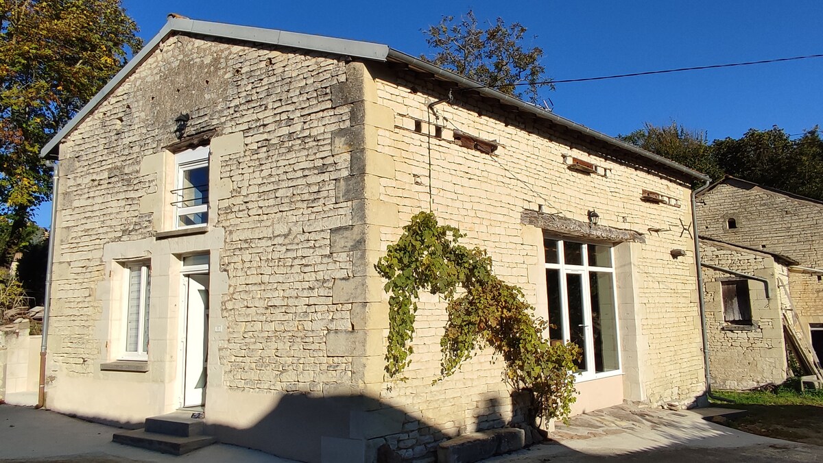 A charming stone exterior of the house is showcased, featuring large windows that allow natural light to fill the interior. Climbing greenery adds a touch of nature, complementing the rustic appearance. The serene blue sky provides a bright backdrop to the inviting façade.