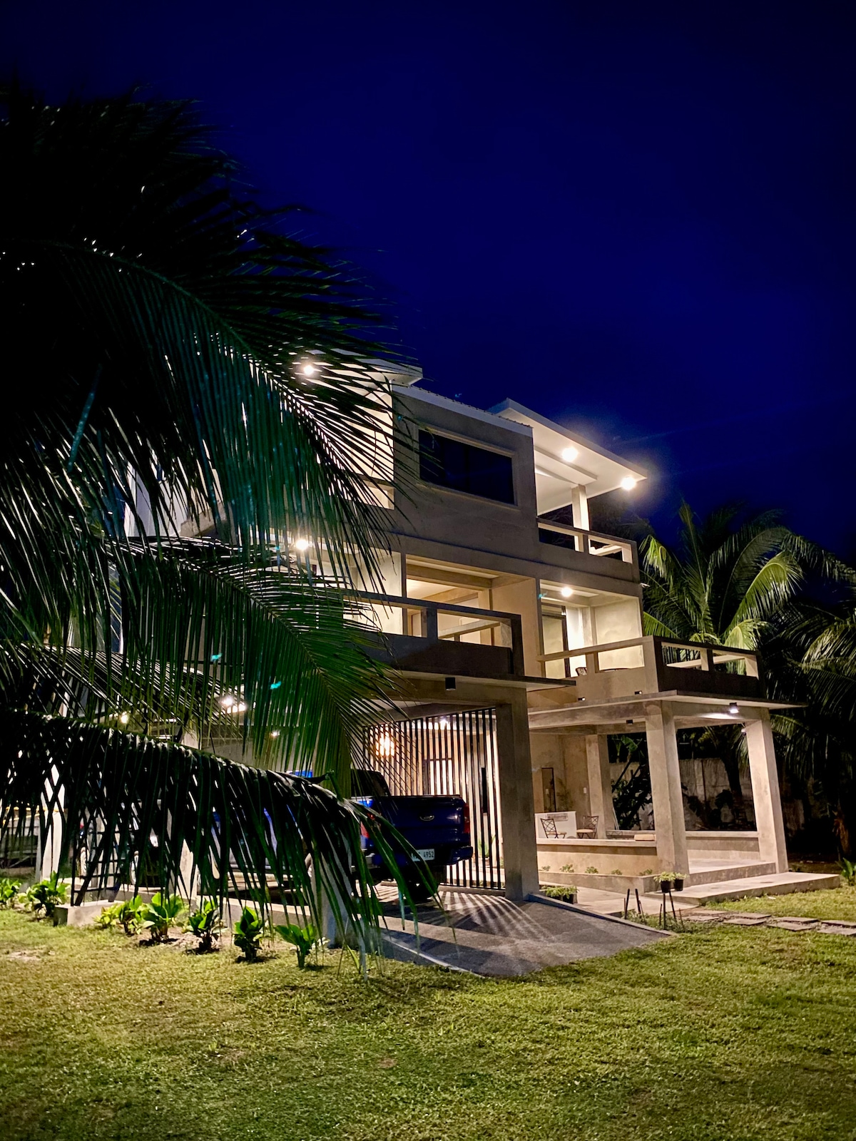A modern two-story building is illuminated under the night sky, highlighting its architectural features. The façade is decorated with warm lighting, creating a welcoming appearance. Surrounding palm trees add to the tropical ambiance, while a manicured lawn enhances the overall setting.
