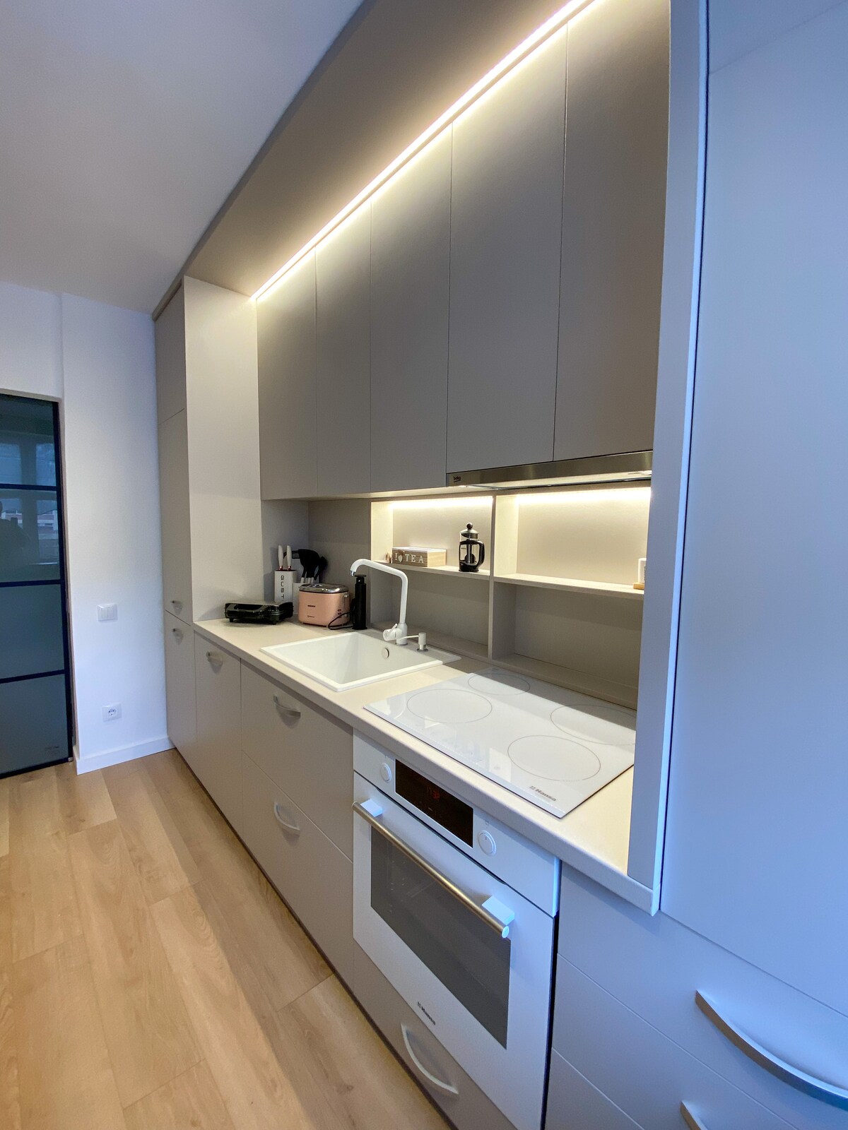 The modern kitchen showcases a sleek design with light-colored cabinetry. Under-cabinet lighting illuminates the countertop, featuring a double sink. A built-in oven and a stovetop are integrated seamlessly into the cabinetry, providing a functional space for meal preparation.
