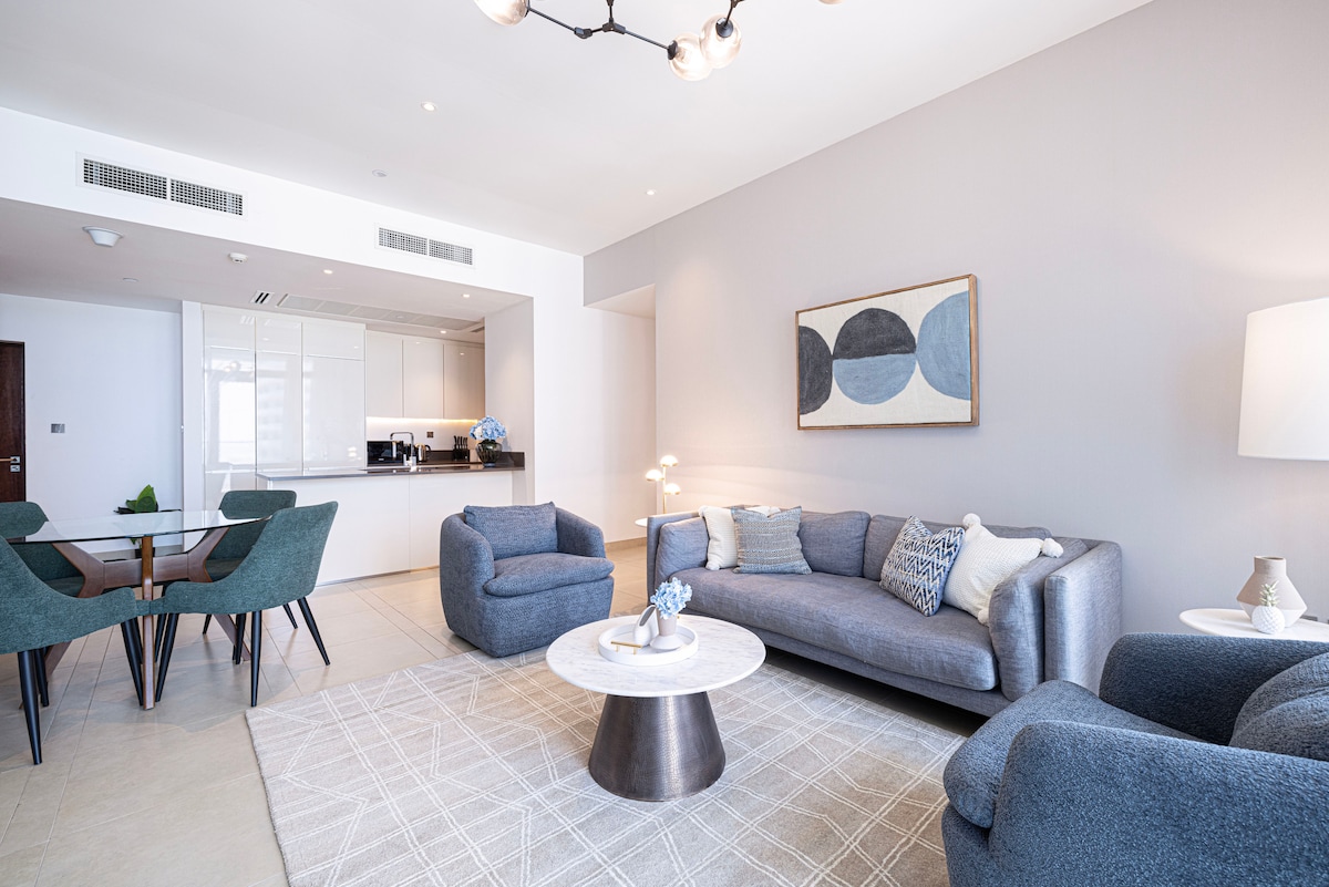 A spacious lounge area features a soft blue sofa and two matching armchairs, arranged around a round marble coffee table. A dining area with four chairs is visible in the background, along with a modern kitchen showcasing sleek cabinetry.