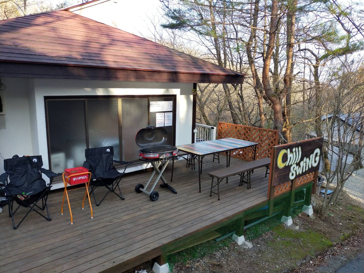 An outdoor deck features a wooden surface with several seating options. Two folding chairs are positioned next to a portable grill, while a larger table with benches occupies the center. A sign reading 'Chill Swing' is displayed prominently at the end of the deck.