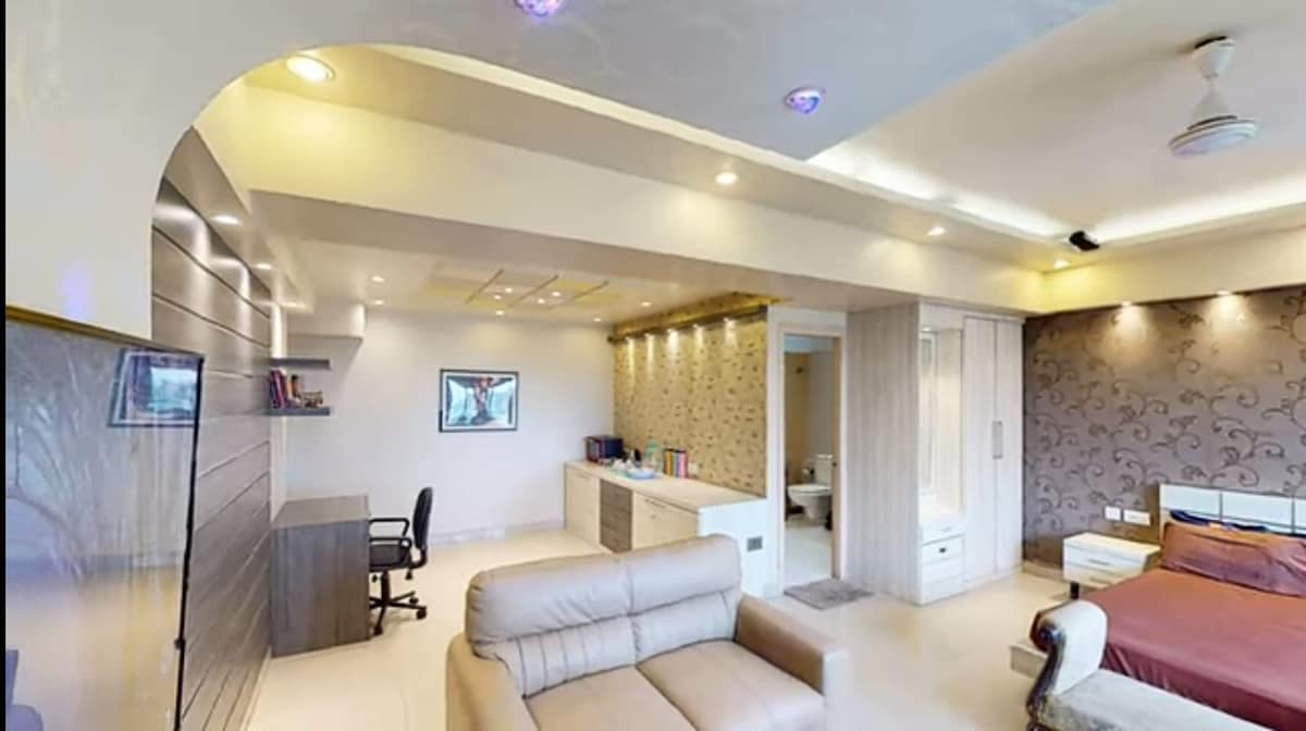 A spacious studio apartment interior features a sleek work desk with a chair, a comfortable sofa, and a king-size bed against a decorative wall. Soft lighting is provided by recessed ceiling lights, highlighting the modern design and open layout towards the en-suite bathroom.