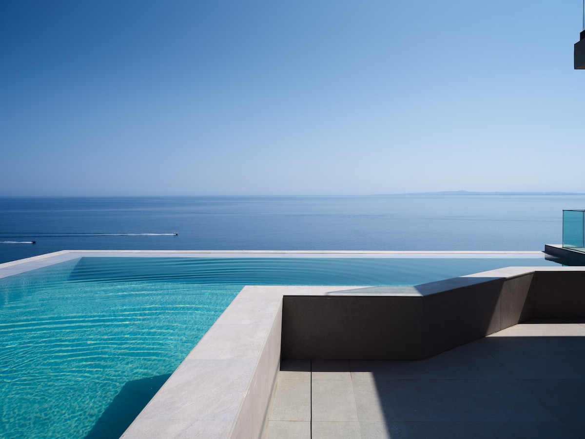 An infinity-edge pool seamlessly blends with the horizon, offering unobstructed views of the vast blue sea. The pool area features smooth, light-colored stone surrounds that enhance the tranquil setting, inviting relaxation and enjoyment of the serene coastal landscape.