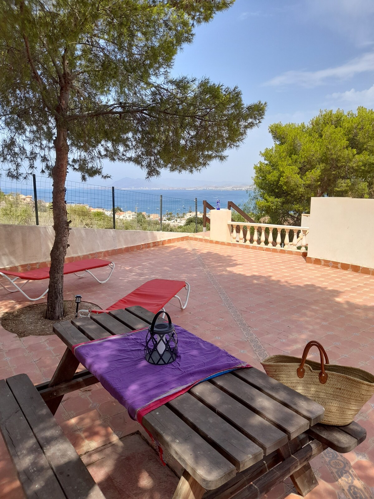 An outdoor dining area features a wooden table set with a purple tablecloth and a decorative lantern. Nearby, two sleek lounge chairs are positioned for relaxation, with views of the water and surrounding greenery visible beyond. A woven basket rests on the table.