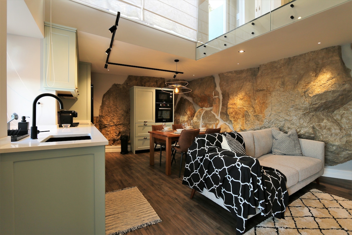 A living area features natural stone walls complemented by modern furnishings. A spacious sofa is adorned with patterned throws, and a dining table is set for meals. Overhead lighting and open spaces enhance the airy atmosphere, while the kitchen is equipped with contemporary appliances.