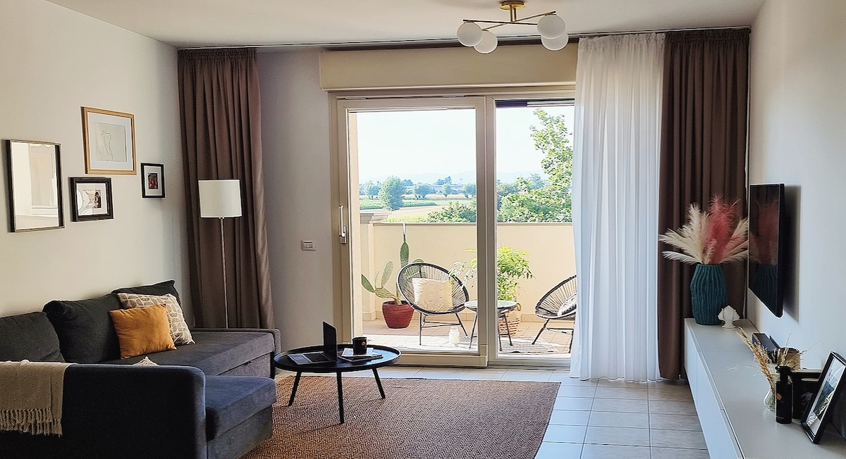 A bright living area is featured, showing a comfortable sofa with decorative cushions and a coffee table. Large glass doors open to a balcony, revealing a view of greenery. Natural light filters in through sheer and dark curtains, enhancing the relaxing ambiance.