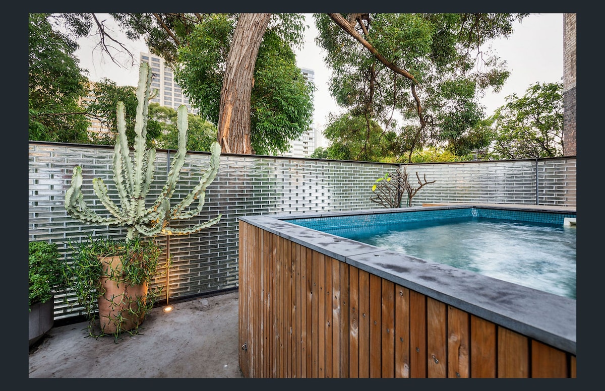 An outdoor entertaining space features a wooden deck surrounding a plunge pool, which is currently under maintenance. A large cactus plant and various leafy greens add natural elements, while a structured glass wall provides privacy and a clear view of the surrounding trees.