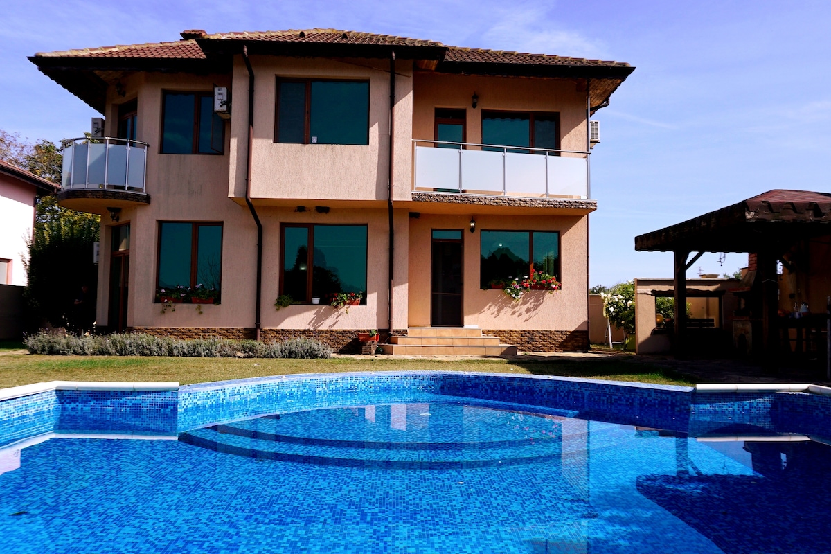The exterior of a two-story villa is displayed, featuring large windows and balconies adorned with flowers. A private swimming pool, with crystal-clear blue water, reflects the villa's structure and is surrounded by well-maintained grass and a covered gazebo.