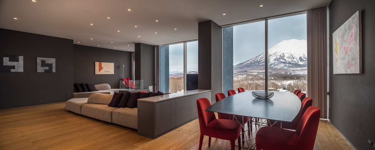 An open-concept living area features large picture windows offering views of Mt. Yotei. Contemporary furnishings include a spacious sofa and a dining table surrounded by red chairs. Neutral walls and wood flooring provide a modern backdrop, while natural light enhances the ambience.